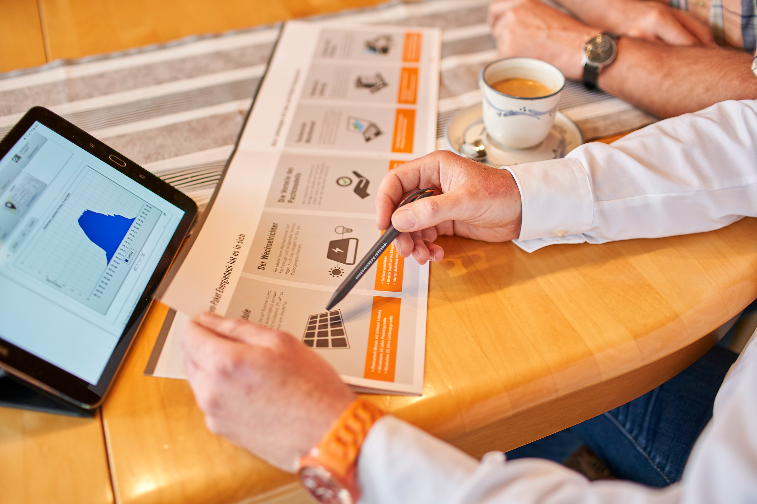 PV-Anlage in Bendorf Ein Tablet zeigt ein Diagramm, während eine Person mit Kugelschreiber ein Prospekt studiert. Eine zweite Person hält eine Tasse Kaffee. Beide sitzen an einem Holztisch.