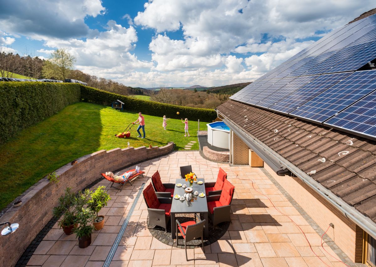 Ein Garten mit grünem Rasen. Eine Person mäht den Rasen, während zwei Kinder spielen. Auf der Terrasse stehen ein Tisch und Stühle, daneben ein Haus mit Solarpanelen auf dem Dach.
