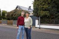 Ein Mann und eine Frau stehen vor einem Haus mit einer Solaranlage und lächeln freundlich.