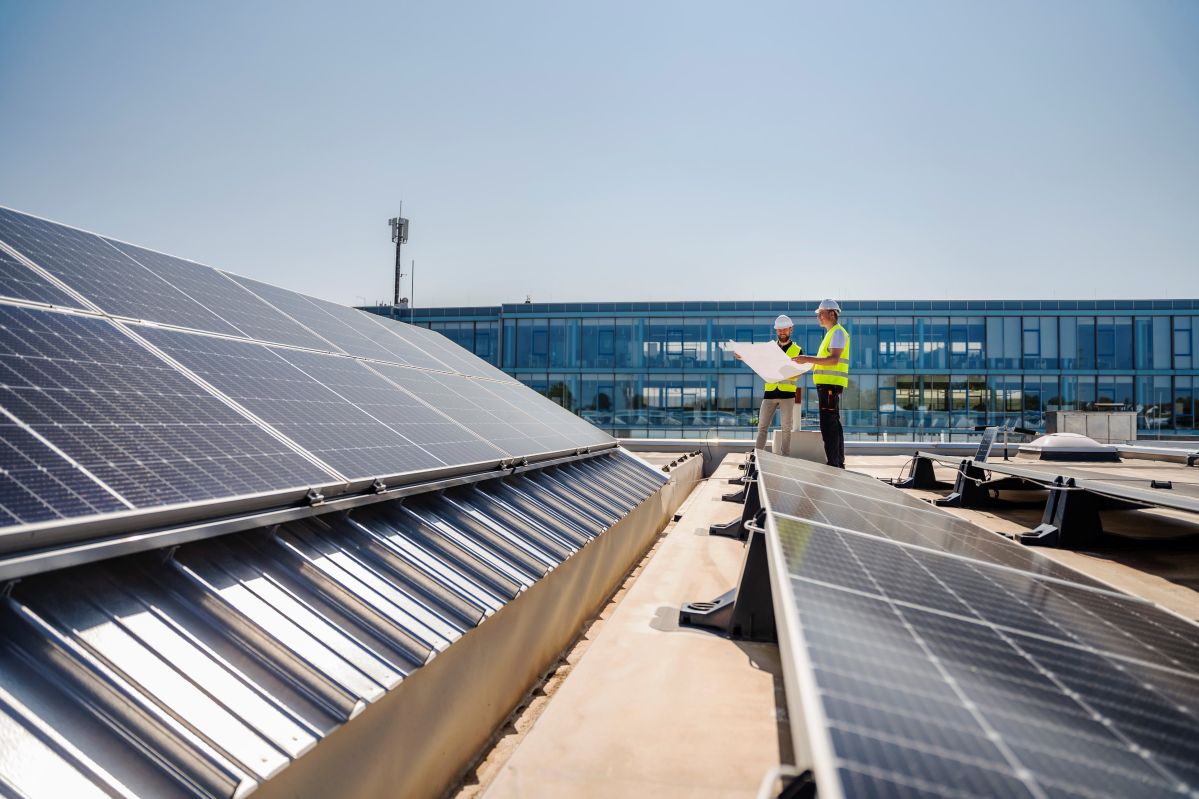 Zwei Männer mit gelben Schutzwesten und Schutzhelmen stehen auf einem Firmendach mit Solarpanelen.