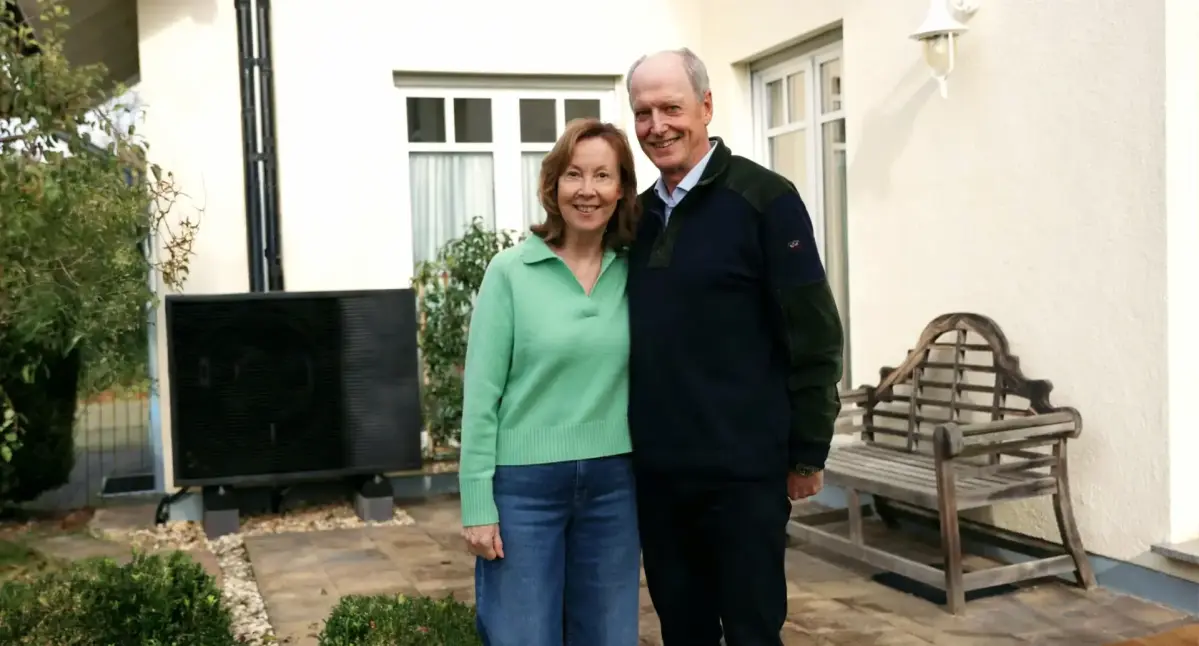 Ein lächelndes Paar steht Arm in Arm auf einer Terrasse vor einem hellen Gebäude mit Fenstern. Neben ihnen befindet sich eine Wärmepumpe und ein schwarzes Schild, umgeben von Pflanzen.