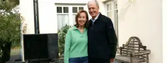 Ein lächelndes Paar steht Arm in Arm auf einer Terrasse vor einem hellen Gebäude mit Fenstern. Neben ihnen befindet sich eine Wärmepumpe und ein schwarzes Schild, umgeben von Pflanzen.