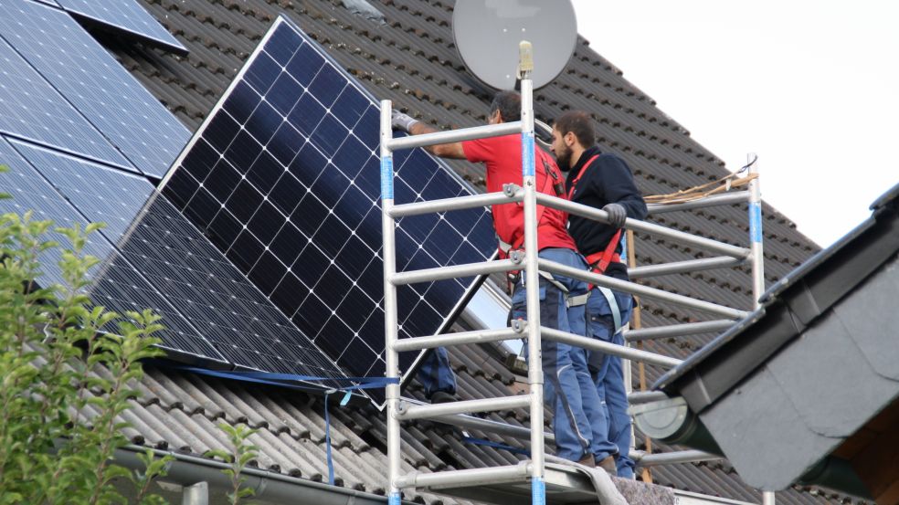 Ein Solarpanel wird auf einem Dach installiert von zwei Männern in Arbeitskleidung. Sie stehen auf einem Gerüst.