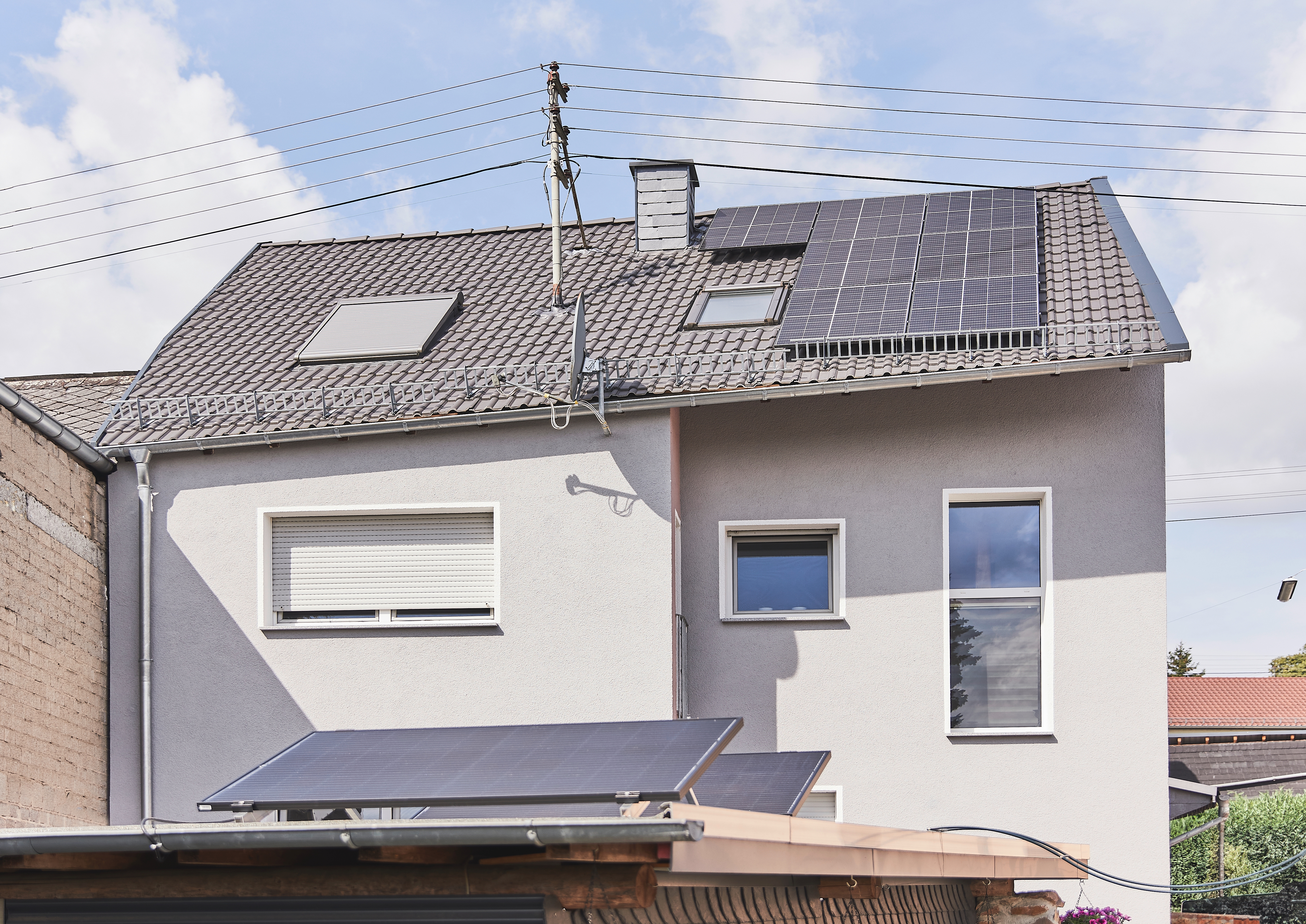 Dein PV-Lexikon Ein zweistöckiges Haus mit Solarpanelen auf dem Dach und einem angrenzenden Flachdach. Der Himmel ist blau, und im Hintergrund sind einige Stromleitungen sichtbar.