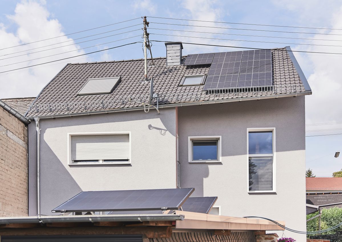 Dein PV-Lexikon Ein zweistöckiges Haus mit Solarpanelen auf dem Dach und einem angrenzenden Flachdach. Der Himmel ist blau, und im Hintergrund sind einige Stromleitungen sichtbar.