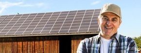 Solaranlage auf Scheune Eine Holzscheune mit Solarpaneelen auf dem Dach. Ein Mann mit gekreuzten Armen und Baseballkappe steht vor der Scheune, die von hellem Tageslicht umgeben ist.