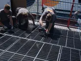 Arbeitskräfte installieren Solarmodule auf einem Dach, verwenden Werkzeuge und tragen Handschuhe im Freien bei sonnigem Wetter, abgesichert durch ein blaues Schutznetz.