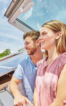 Ein Paar steht in einem Dachfenster und schauen raus. Auf dem Dach befindet sich eine Solaranlage.