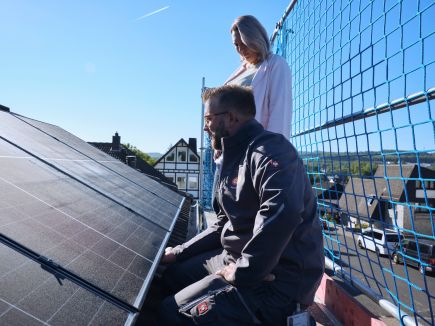 Eine evm Handwerker und eine Frau sind auf einem Baugerüst. Sie stehen bzw. knien neben einer Solaranlage auf einem Dach.