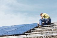 Ein Mann montiert Solarpaneele auf einem Dach. Er trägt Arbeitskleidung und ist bei klarem Himmel über einer Ziegeldachstruktur beschäftigt.