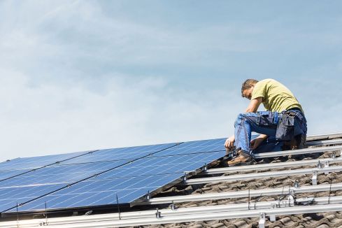 Ein Mann montiert Solarpaneele auf einem Dach. Er trägt Arbeitskleidung und ist bei klarem Himmel über einer Ziegeldachstruktur beschäftigt.