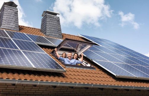 Eine Familie steht in einem Dachfenster und schaut fröhlich raus. Auf dem Dach befindet sich eine Solaranlage.
