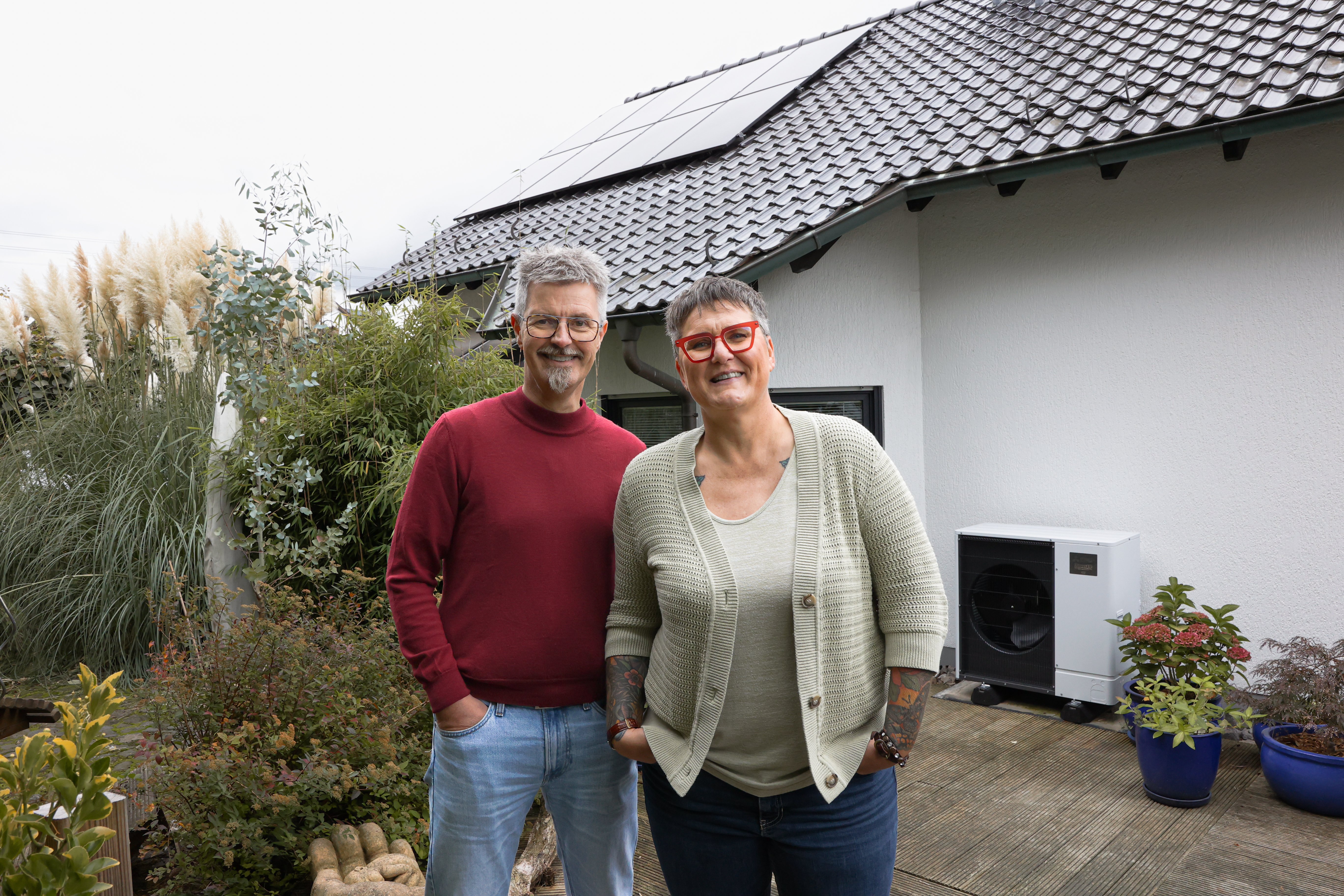 Ein Mann und eine Frau stehen vor ihrem Haus. Im Hintergrund ist eine PV-Anlage und eine Wärmepumpe zu sehen.