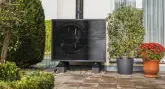 Eine schwarze Wärmepumpe steht stationär auf einem gepflasterten Bereich vor einer Hauswand, umgeben von grünen Büschen und blühenden Pflanzen in Töpfen.