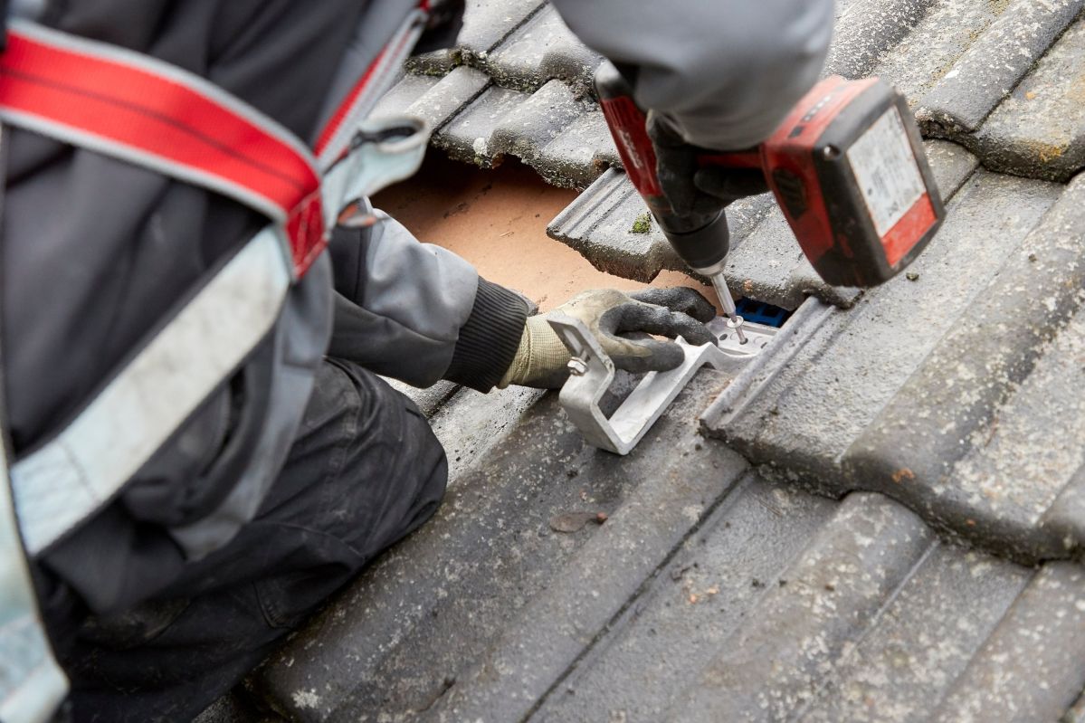 Eine Person repariert ein Loch in einem Schieferdach mit einer Bohrmaschine. Umgeben von grauen Dachziegeln, trägt die Person Arbeitshandschuhe und eine Sicherheitsweste.