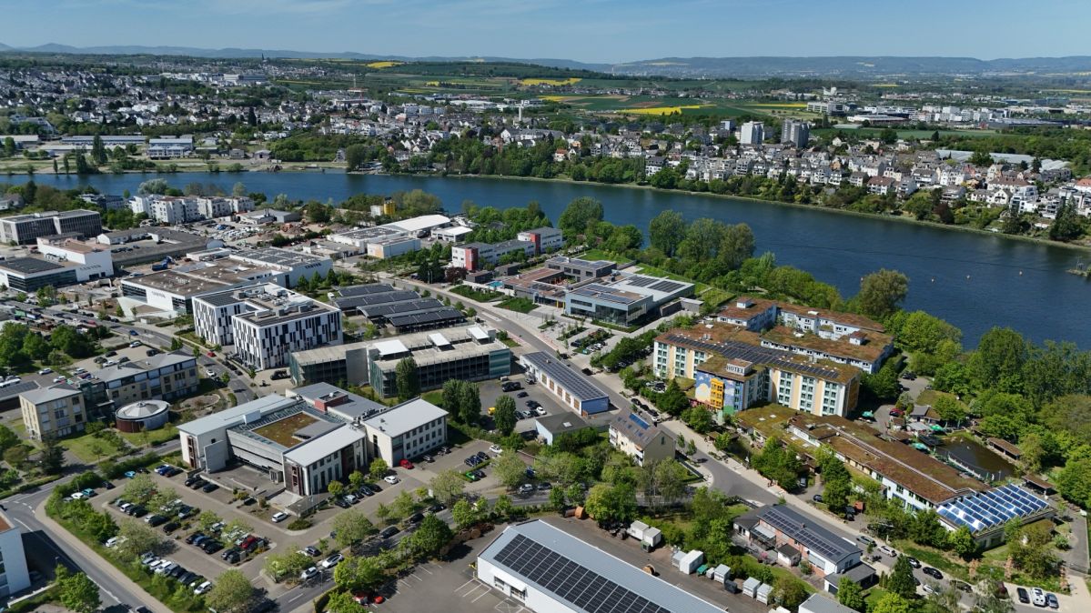 Luftaufnahme eines Gewerbe- und Bürostandorts am Rheinufer mit modernen Gebäuden und angrenzender Wohnbebauung.