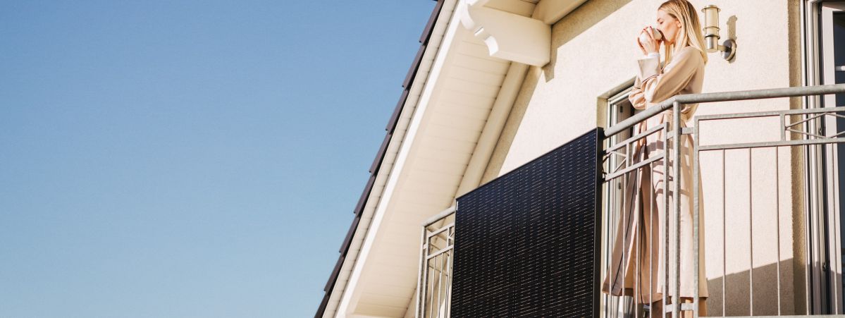 Ein Solarpanel ist an einem Balkongelände montiert. Eine Frau steht auf dem Balkon und trinkt aus einem Becher.