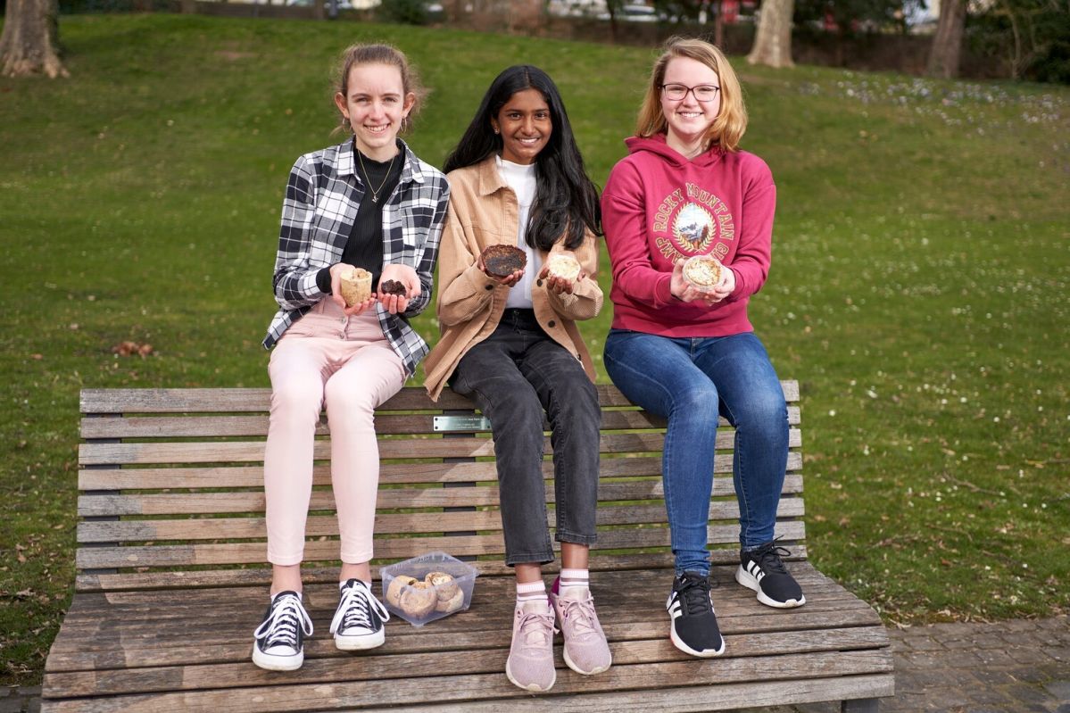 Drei Personen sitzen auf einer Parkbank und halten jeweils Apfelteller in den Händen. Im Hintergrund sind Wiese und Bäume zu sehen. Ein Beutel mit weiteren Apfeltellern liegt daneben.