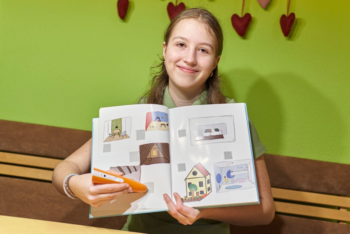 evm magazin Eine Person hält ein geöffnetes Buch mit bunten Illustrationen, während sie an einem Tisch sitzt. Im Hintergrund sind grüne Wände mit dekorativen Herzanhängern zu sehen.