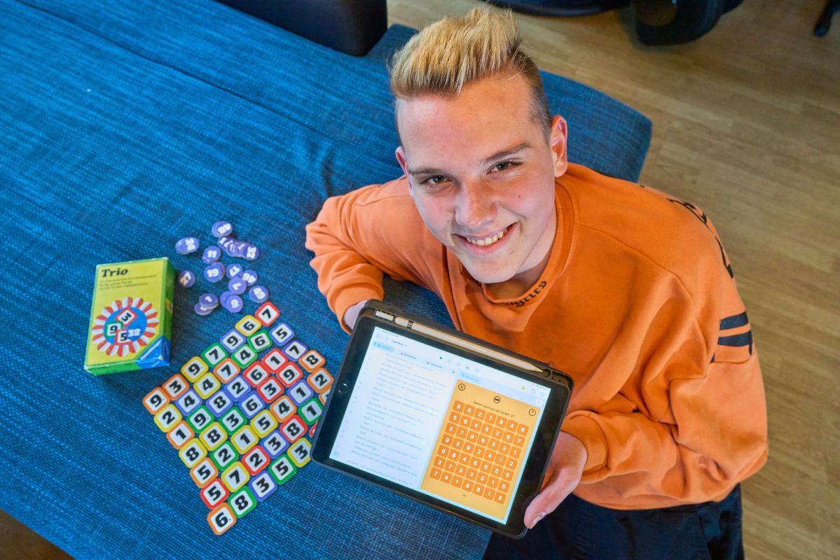 Eine Person sitzt an einem Tisch, hält ein Tablet mit einem Spielgitter, daneben liegen das Spiel "Trio", Zahlenchips und eine Spielanleitung liegen auf einer blauen Tischdecke.