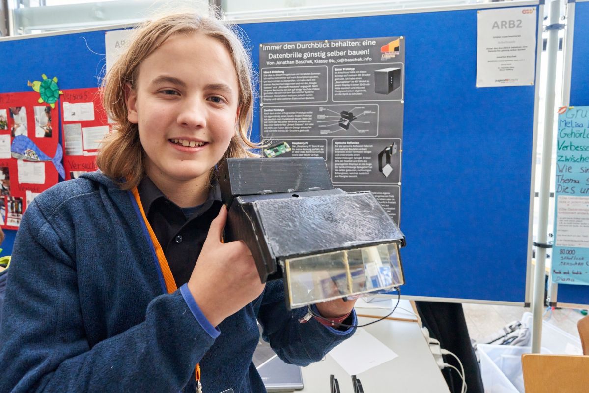 Eine Person hält ein selbstgebautes elektronisches Gerät vor einer Präsentationstafel in einer Ausstellungshalle. Auf der Tafel steht: "Innen den Durchblick behalten: eine Datenbrille günstig selber bauen".