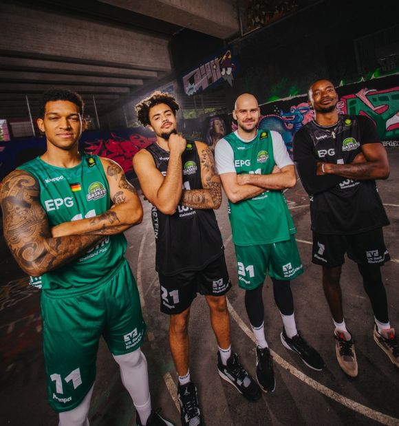 EPG Baskets Vier Spieler der EPG Baskets Koblenz in Teamkleidung stehen auf einem Basketballfeld unter einer Brücke vor einer mit Graffiti gestalteten Wand.