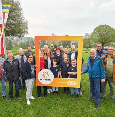 Eine Gruppe lächelnder Menschen steht draußen auf einer Wiese. Einige halten einen großen, orangefarbenen Rahmen mit der Aufschrift "Ehrensache" und "evm". Im Hintergrund sind Bäume und Fahnen zu sehen.