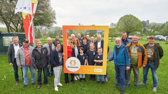 #evm #ehrensache in der VG Wallmerod Eine Gruppe lächelnder Menschen steht draußen auf einer Wiese. Einige halten einen großen, orangefarbenen Rahmen mit der Aufschrift "Ehrensache" und "evm". Im Hintergrund sind Bäume und Fahnen zu sehen.