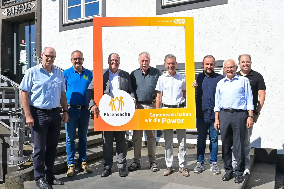 Acht Männer stehen vor dem Eingang eines Rathauses und halten einen orangenen Rahmen mit der Aufschrift "evm Ehrensache". Sie lächeln freundlich.