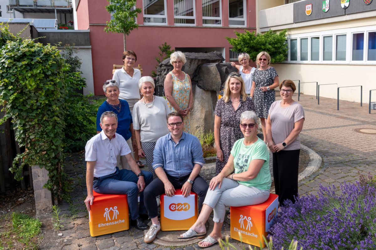 Mehrere Personen stehen vor einem Brunnen und lächeln freundlich. Drei Personen sitze im Vordergrund auf orangenen evm Würfeln mit der Aufschrift "Ehrensache".
