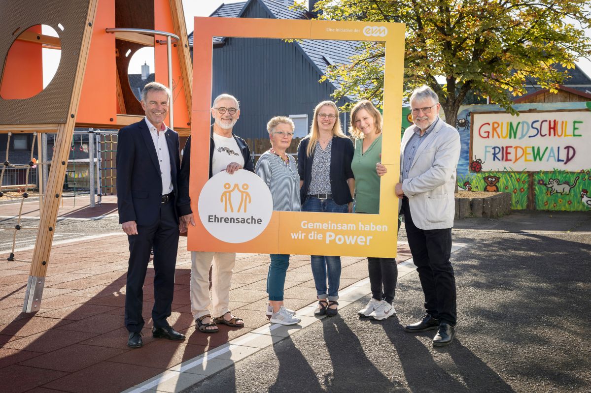 Drei Männer und drei Frauen stehen auf einem Spielplatz und halten einen orangenen Rahmen mit der Aufschrift "evm Ehrensache". Sie lächeln freundlich.