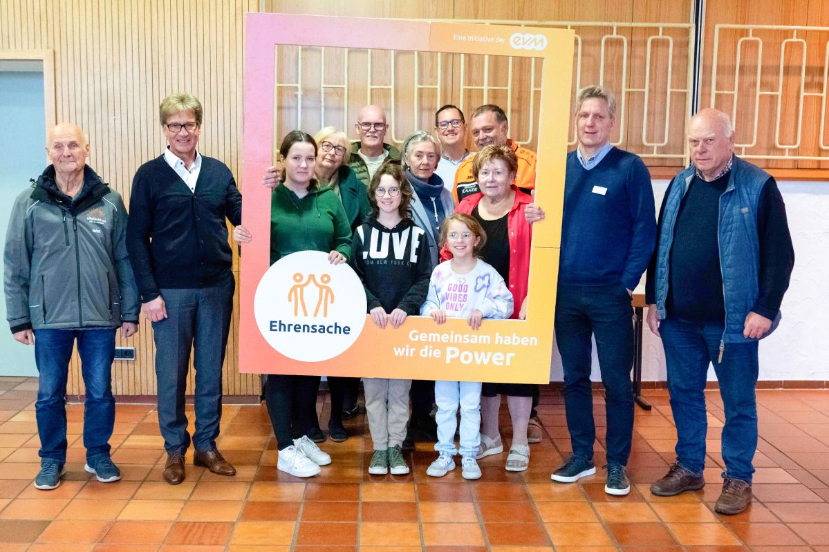 Eine Gruppe von Erwachsenen und Kindern stehen in einem Gebäude und halten einen orangenen Rahmen mit der Aufschrift "evm Ehrensache". Sie lächeln freundlich.