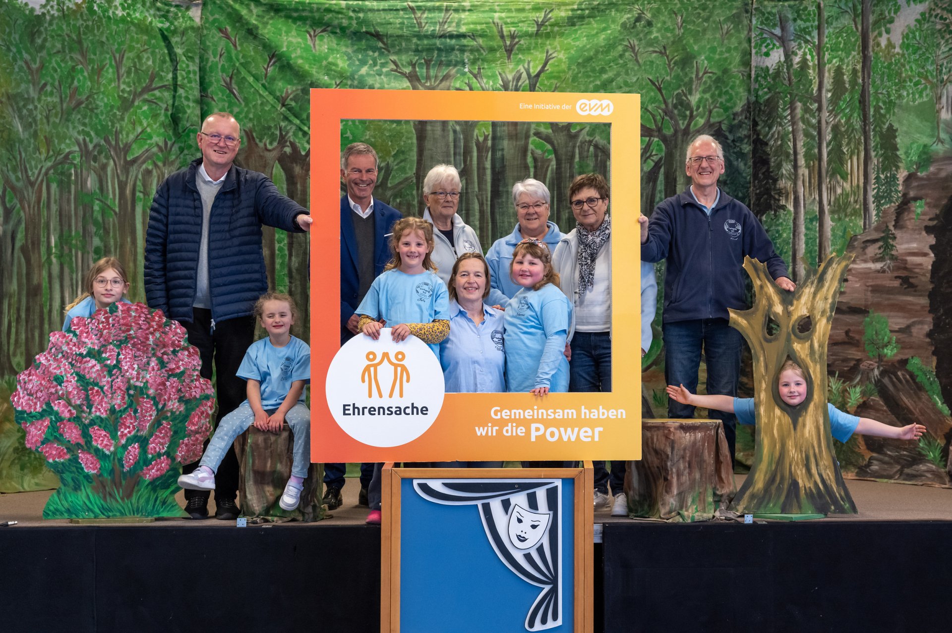 Eine Gruppe von Erwachsenen und Kindern stehen auf einer Theaterbühne und halten einen orangenen Rahmen mit der Aufschrift "Ehrensache".