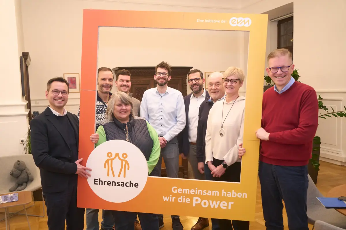 Sieben Männer und zwei Frauen stehen in einem Raum und halten einen orangenen Rahmen mit der Aufschrift "evm Ehrensache". Sie lächeln freundlich.