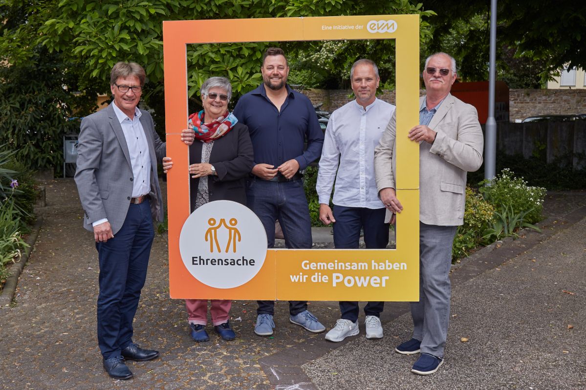 Vier Männer und eine Frau lächeln freundlich und halten einen orangenen Rahmen mit der Aufschrift "Ehrensache".