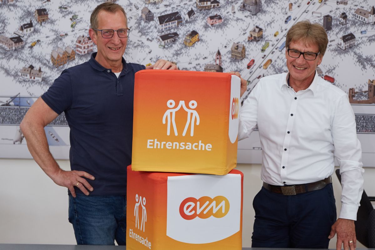 Zwei Männer stehen neben aufeinander gestapelten Würfeln. Die orangenen Würfel haben die Aufschrift "evm" und "Ehrensache".