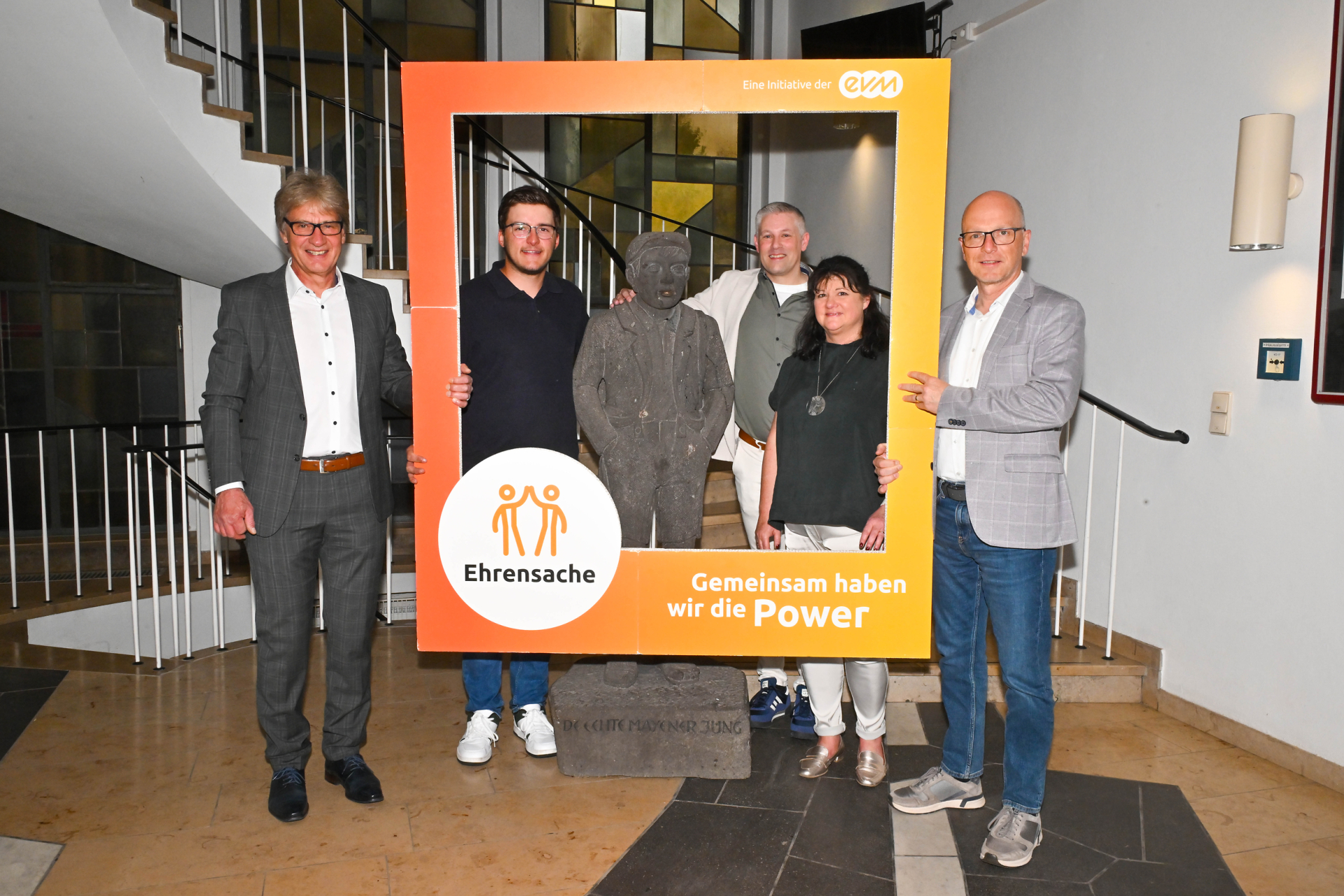 Vier Männer und eine Frau stehen vor einer Statue und halten einen Orangenen Rahmen. Auf dem Rahmen steht "evm Ehrensache". Sie lächeln freundlich und sind professionell gekleidet.