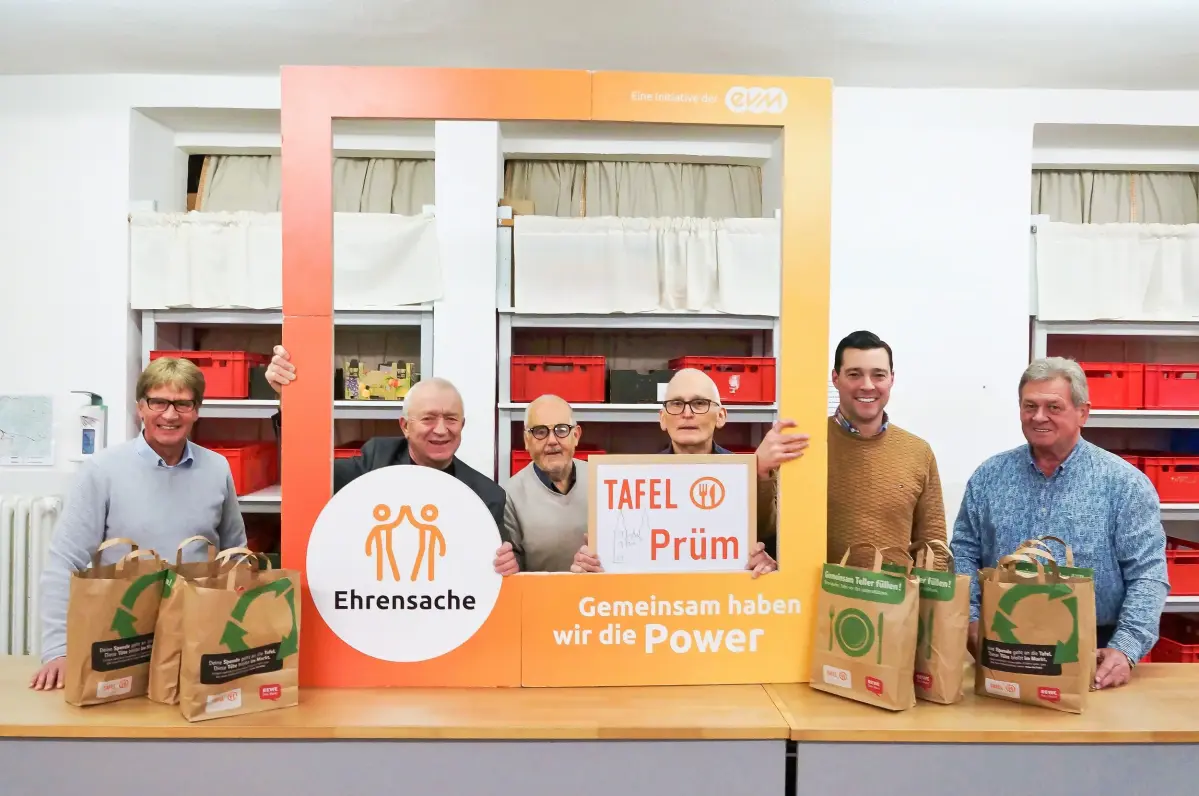 Sechs Männer stehen an einer Ausgabestelle der Tafel und halten einen orangenen Rahmen mit der Aufschrift "evm Ehrensache". Sie lächeln freundlich.