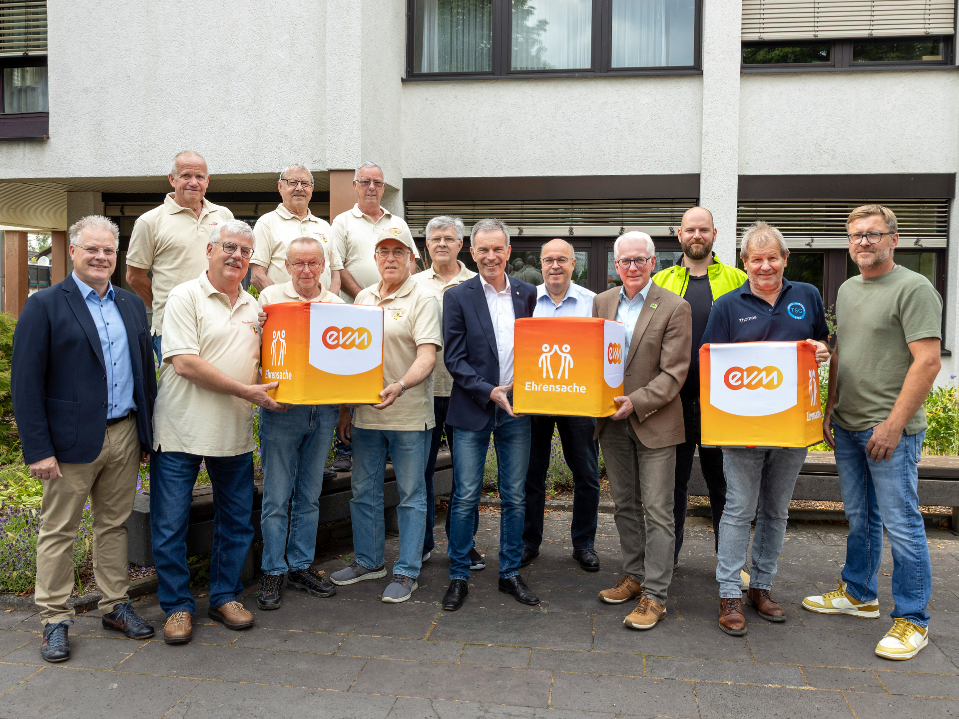 Eine große Gruppe steht vor einem Gebäude. Sie halten orangene Würfel mit der Aufschrift "evm Ehrensache" und lächeln freundlich.