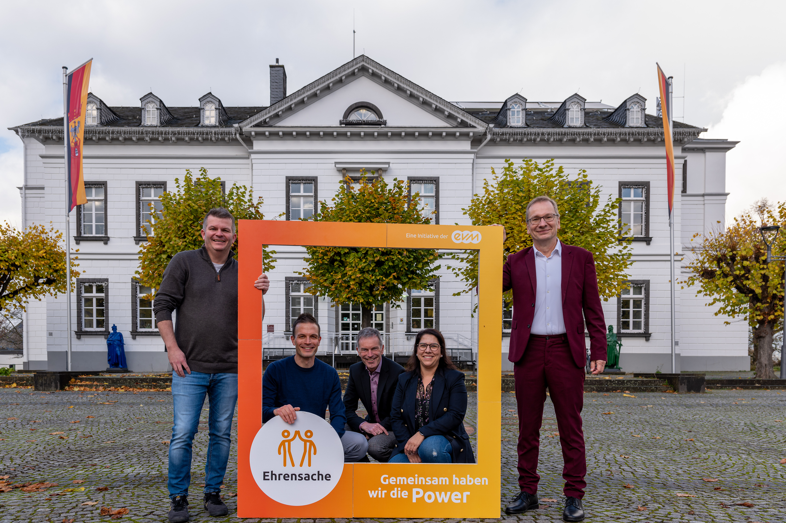 Vier Männer und eine Frau stehen vor einem Amtsgebäude und halten einen orangenen Rahmen mit der Aufschrift "evm Ehrensache". Sie lächeln freundlich.