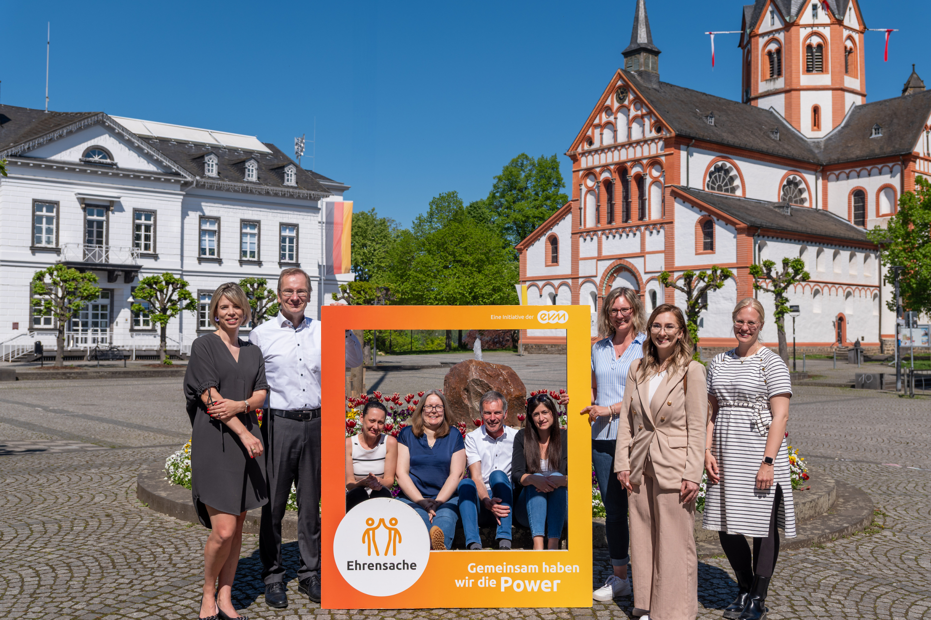 Sieben Frauen und zwei Männer stehen bzw. sitzen auf einem Platz und halten einen orangenen Rahmen mit der Aufschrift "evm Ehrensache". Sie lächeln freundlich. Im Hintergrund ist ein Gebäude und eine Kirche zu sehen.