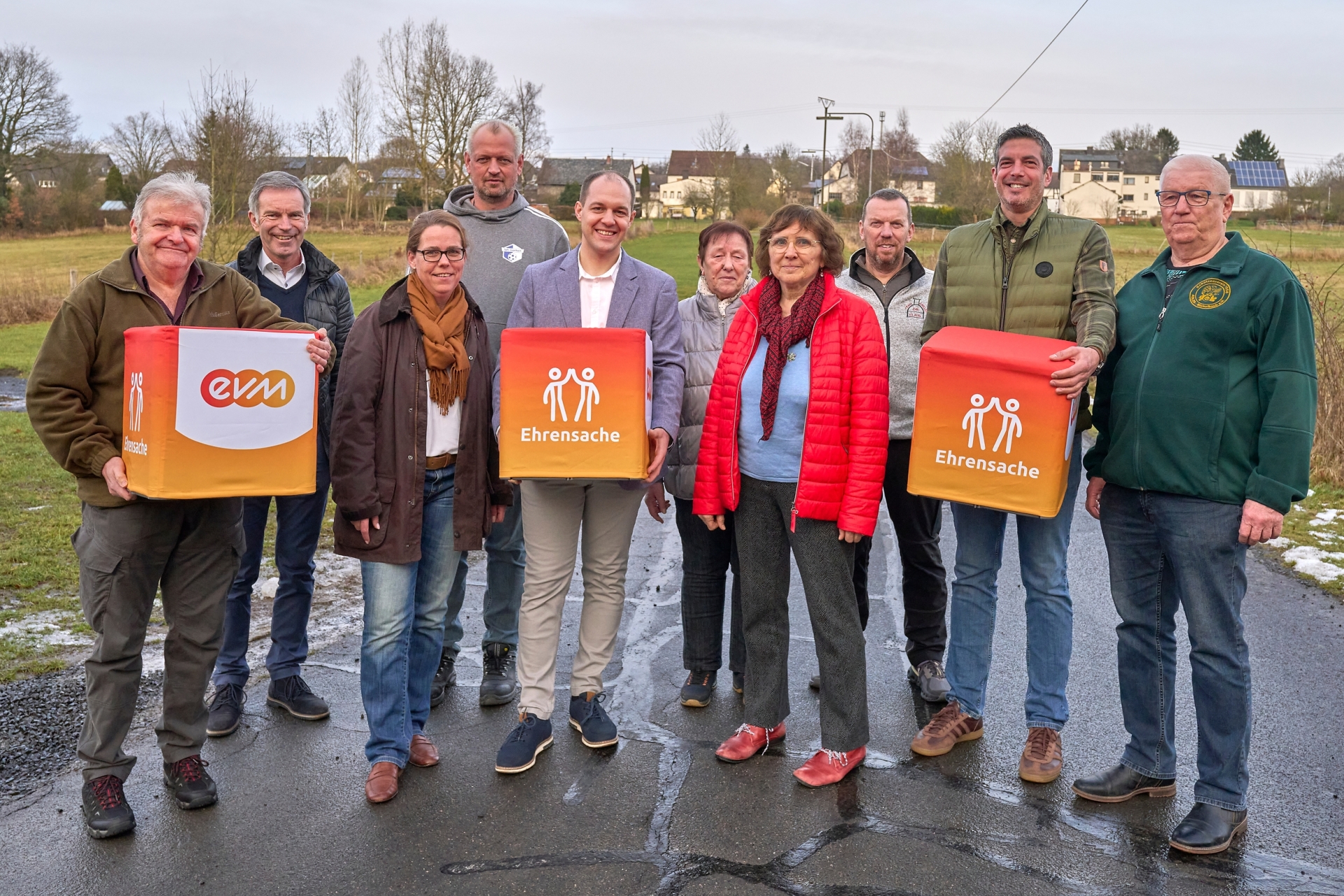 Gruppe von Personen bei einer Spendenübergabe im Rahmen des evm‑Programms ‚Ehrensache‘; mehrere halten orangefarbene Spendenboxen mit evm‑ und Ehrensache‑Logo.