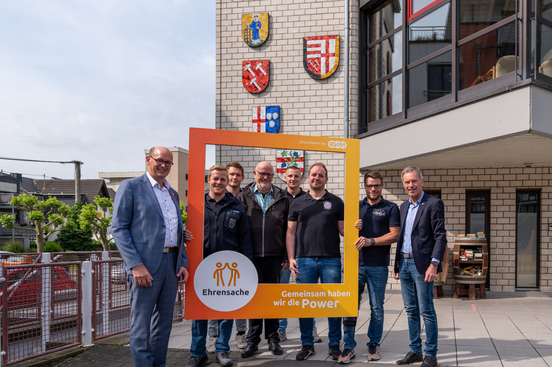 Acht Männer stehen vor einem Gebäude mit Wappen und lächeln freundlich. Sie halten einen orangenen Rahmen mit der Aufschrift "Ehrensache".
