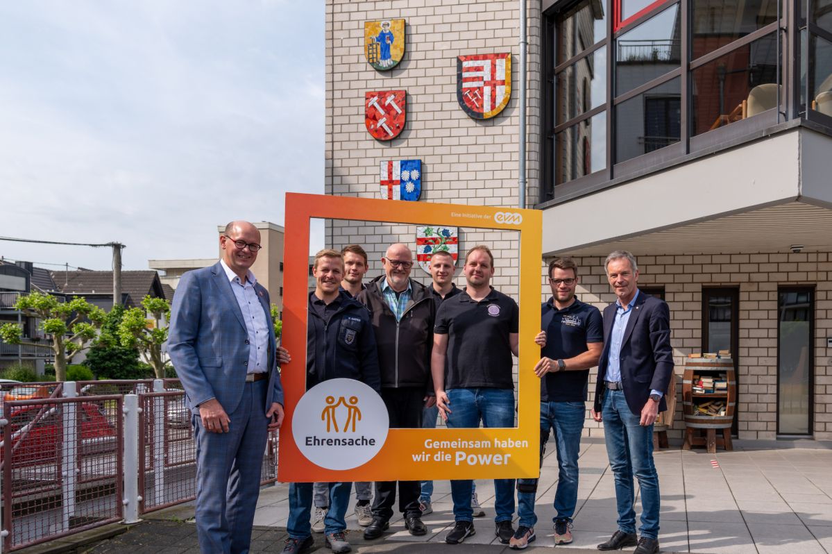 Acht Männer stehen vor einem Gebäude mit Wappen und lächeln freundlich. Sie halten einen orangenen Rahmen mit der Aufschrift "Ehrensache".