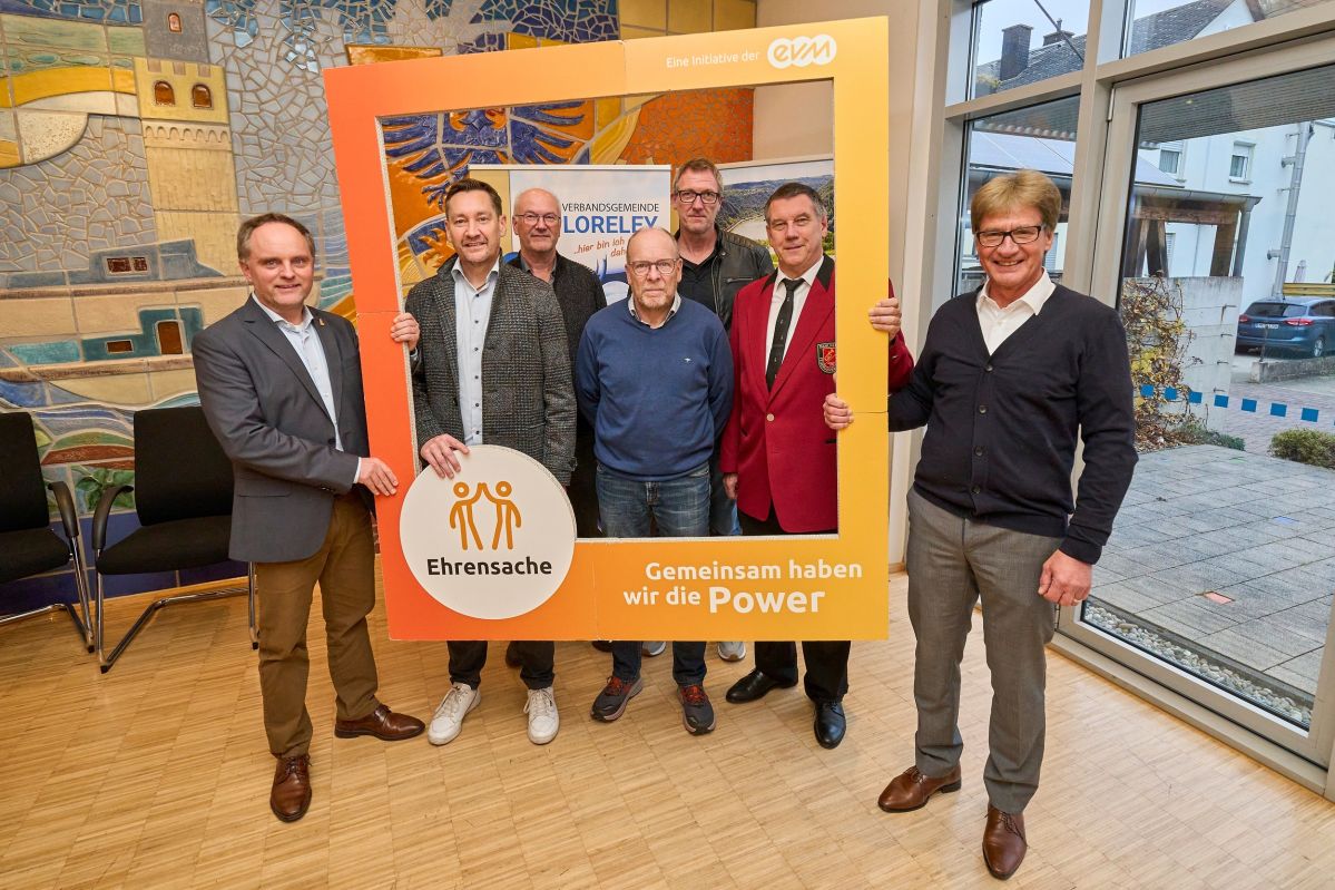 Sieben Männer stehen in einem Raum und halten einen orangenen Rahmen mit der Aufschrift "evm Ehrensache". Sie lächeln freundlich.