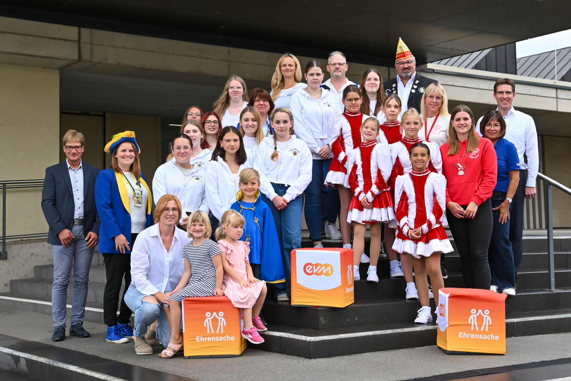 Eine Gruppe an kostümierten Karnevalisten steht vor einem Gebäude-Eingang. Vor ihnen stehen orangene Würfel mit der Aufschrift "evm Ehrensache".
