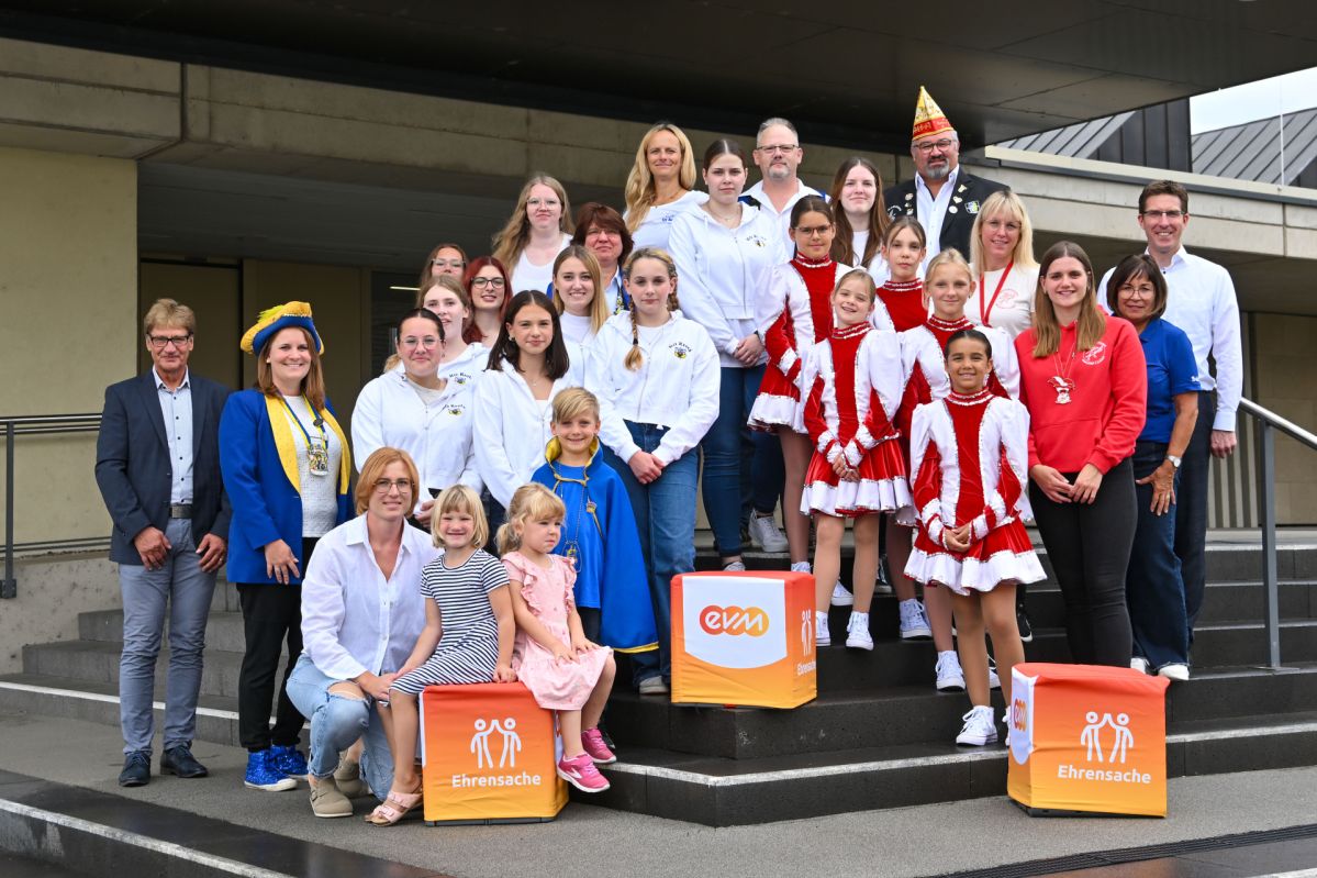 Eine Gruppe an kostümierten Karnevalisten steht vor einem Gebäude-Eingang. Vor ihnen stehen orangene Würfel mit der Aufschrift "evm Ehrensache".