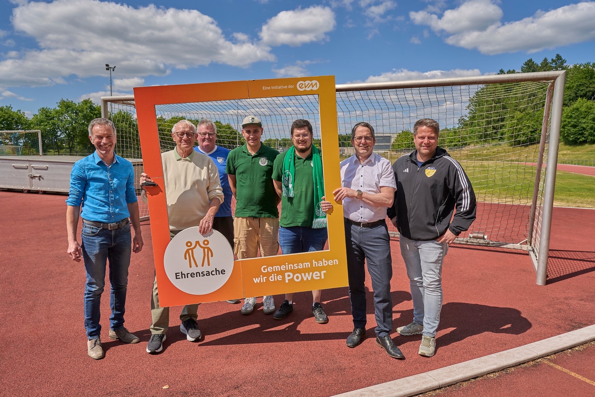 Sieben Männer stehen auf einem Sportplatz und lächeln freundlich. Sie halten einen orangenen Rahmen mit der Aufschrift "Ehrensache".