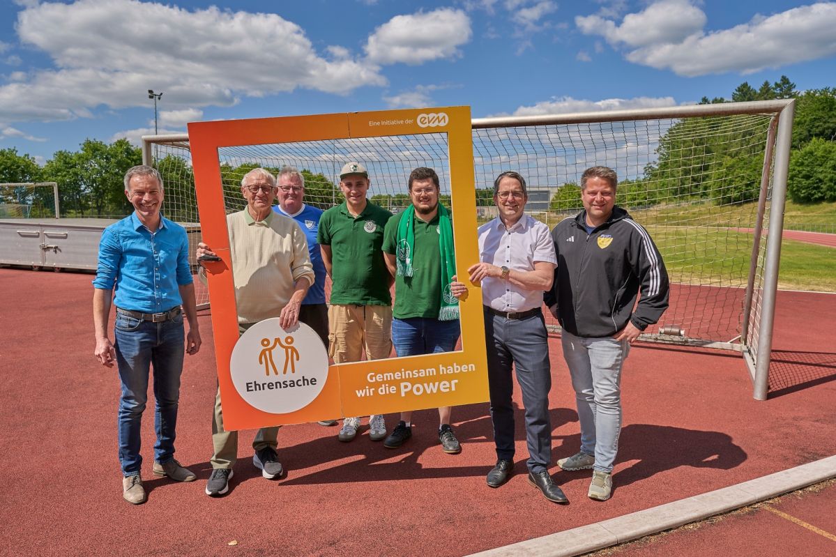 Sieben Männer stehen auf einem Sportplatz und lächeln freundlich. Sie halten einen orangenen Rahmen mit der Aufschrift "Ehrensache".