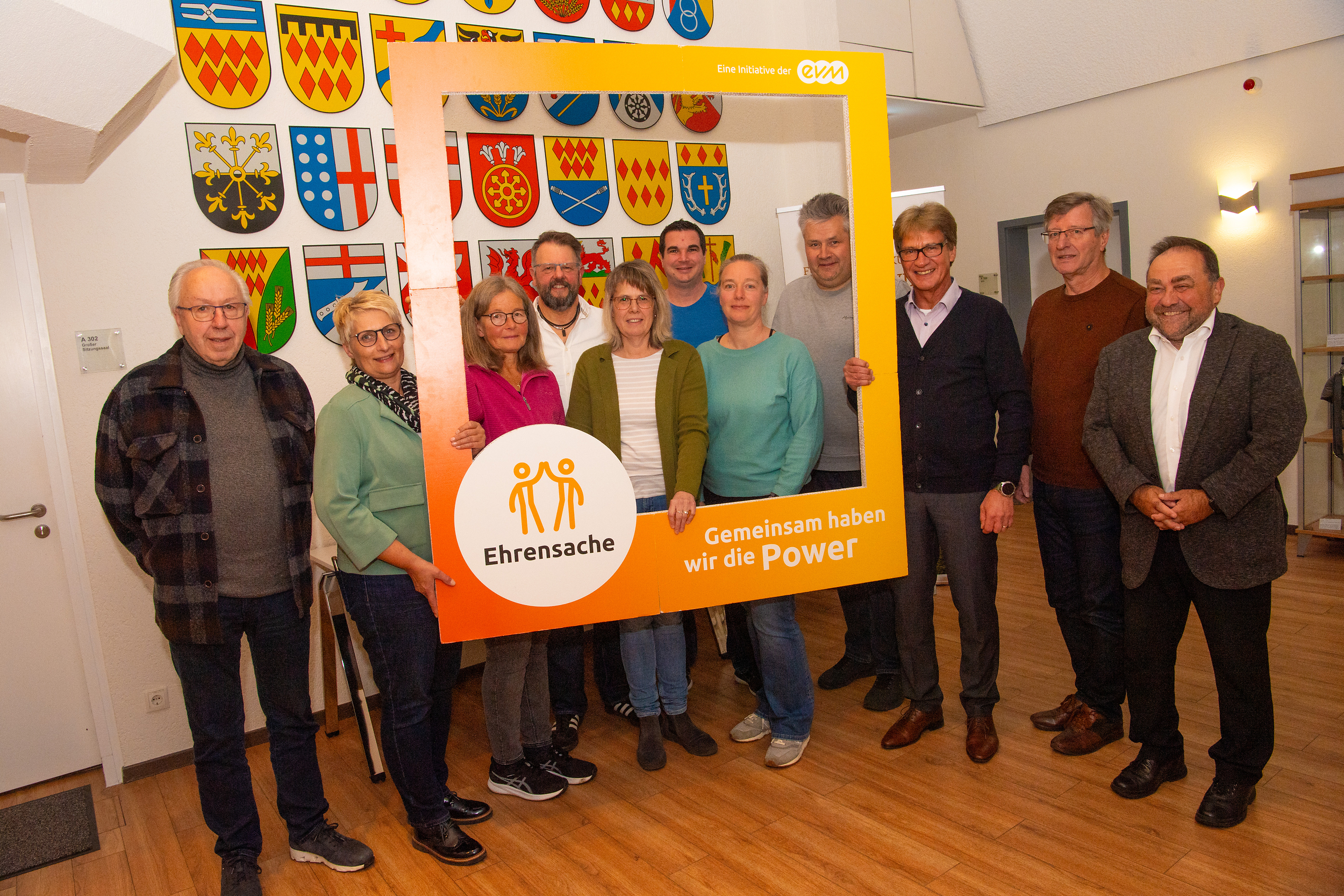 Sieben Männer und vier Frauen stehen in einem Raum und halten einen orangenen Rahmen mit der Aufschrift "evm Ehrensache". Sie lächeln freundlich.